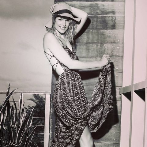 black and white photo of a woman with blonde hair wearing a sun hat and a long patterened backless dress holding her hat and the end of her dress up in front of a building