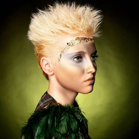 woman with short blonde spiked up hair looking in the distance to the right wearing a flanel with green feathers attached to her feather in front of a green gradient background