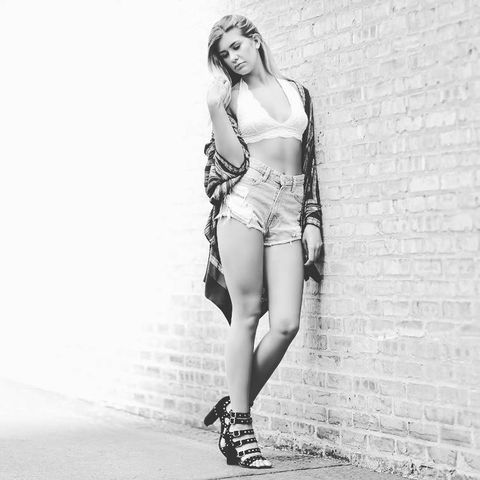 photo in black and white of a woman with long light colored hair wearing a sports bra, short jeans and an open long flanel  and heels leaning up against a brick wall