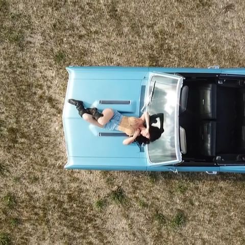woman in a yellow top, short jean shorts, and tall black boots laying on the hood of a light blue convertible muscle car
