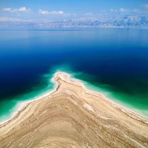 Landscape of a sandy beachside that comes to a point in dark blue water with mountains seen off in the distance under a blue semi-cloudy sky