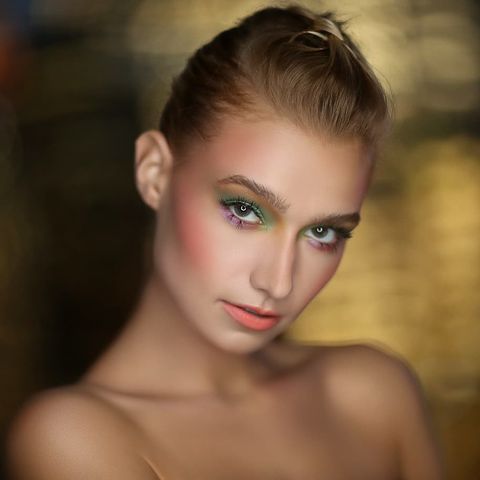 Woman with pulled back blonde hair and green eyes with her head tilted to the side standing in front of an out of focus yellow background