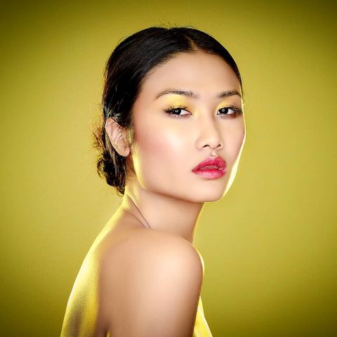 Woman with long dark pulled back hair and dark brown eyes with her mouth very slightly open standing towards the right with her head tilted towards the camera seen from the bare shoulder up in front of a dark yellow gradient background